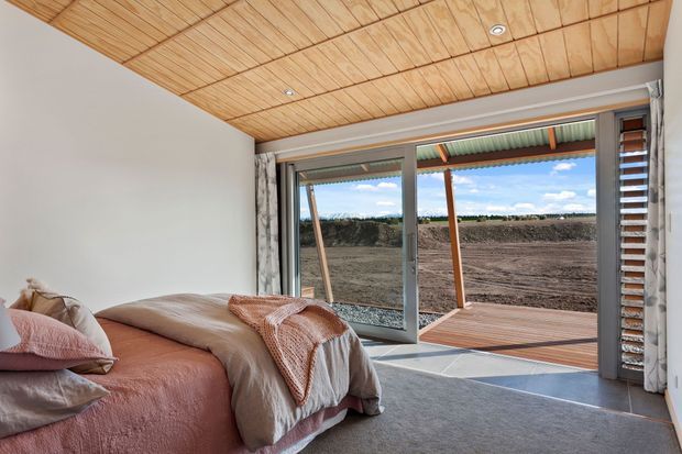 A bright bedroom with a low-pitched timber-lined ceiling, soft neutral and blush bedding, grey carpet, and full-height sliding glass doors opening to a covered timber deck. External horizontal louvres provide sun control, with an open rural landscape visible beyond.
