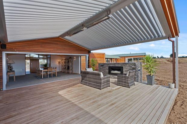 A covered outdoor entertaining deck with a large adjustable louvre roof, timber-clad walls, built-in outdoor fireplace, and wicker seating. The space opens directly to an indoor dining and living area, with a dry landscape beyond.