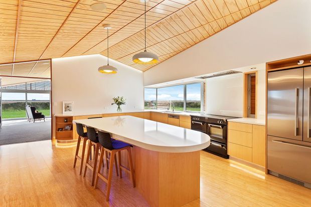 A bright open-plan kitchen with a large curved white island and integrated seating, extensive timber cabinetry, stainless-steel appliances, and floor-to-ceiling glazing. The room features a distinctive warm timber-lined sloping ceiling with pendant lights and connects visually to a living area and outdoor landscape.