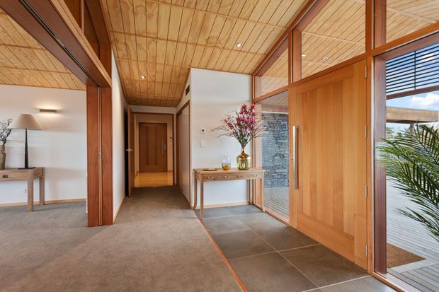 Warm timber-lined residential entry hall with a carpeted living area to the left, a tiled foyer, timber-framed glazing and a large timber pivot door. A slim console, floral arrangement, exposed stone feature wall and glimpses of an exterior courtyard add visual interest. The space has a strong mid-century-inspired architectural character with extensive natural timber detailing.