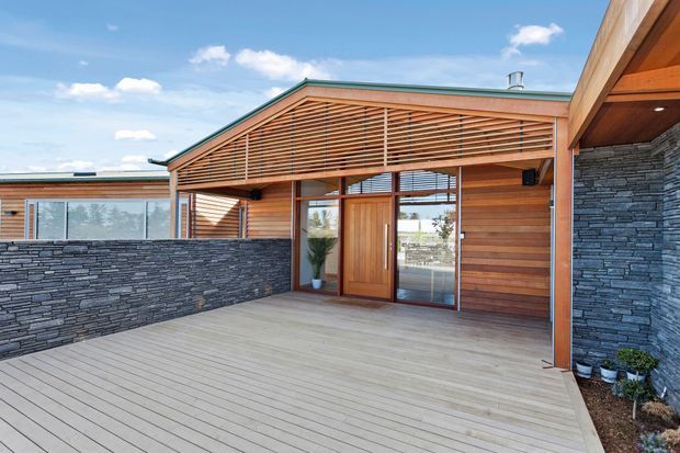 A contemporary single-storey home entrance and covered outdoor terrace. The facade combines warm horizontal timber cladding and a slatted timber sunshade with dark stacked-stone walling, glazed doors, and a broad timber deck. The low-pitched roof and bright blue sky create an open, welcoming exterior setting.