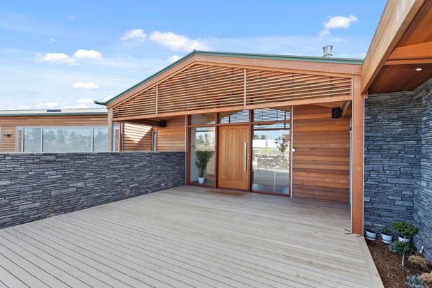 Single-storey contemporary residential exterior with a broad timber-clad entrance pavilion opening onto a spacious timber deck. Horizontal timber screening spans the gabled facade above a timber entry door and glazed sidelights, while dark stacked-stone walls frame the deck and side elevation. The image shows a bright outdoor entry and entertaining area beneath a clear blue sky.