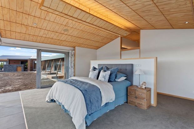 A contemporary bedroom with a large upholstered bed, layered blue and white bedding, and a timber bedside chest. The room features a warm timber-lined ceiling with recessed lighting, grey carpet, white partition walls, and extensive glazing opening toward an exterior courtyard or construction area. A partial open mezzanine or storage zone is visible behind the bed.