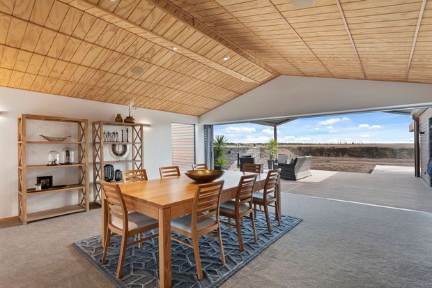 Open-plan dining area with a large solid timber dining table and matching chairs, positioned beneath a distinctive angled timber-panelled ceiling. Built-in or open timber shelving displays crockery and decor along the wall. Full-height corner glazing and sliding doors connect the dining space to a spacious timber deck and outdoor seating area, creating a bright indoor-outdoor connection. The room has neutral carpet, white walls, warm timber detailing, and a relaxed contemporary coastal feel.