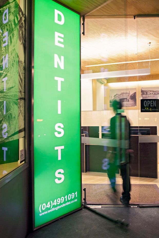 Photograph of a commercial retail or showroom entrance viewed from outside through an open glass door. A large green vertical sign reads “DENTISTS,” with a reception counter and cabinetry visible inside. The interior features warm timber wall and ceiling finishes, dark cabinetry, framed wall graphics, suspended linear lighting, and a person entering the space.