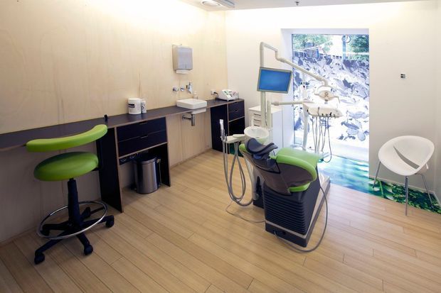 Bright contemporary dental treatment room with a dental chair and overhead examination light, compact clinical cabinetry and sink along the left wall, a wall-mounted monitor, and dental instruments. The room has pale timber-look flooring, light neutral walls, green and white seating accents, and a large window with a decorative patterned view.