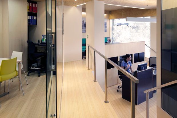 A bright contemporary office interior viewed from an upper-level corridor or landing. The space features pale timber flooring, white walls and partitions, glass office fronts, a reception or workstation area below, built-in desks and shelving, and simple metal handrails around the stair or mezzanine edge. Blue and lime-green chairs add colour, while large windows provide daylight.