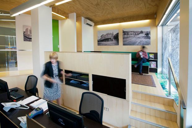 A bright contemporary commercial reception or office interior with a timber-lined ceiling, pale timber built-in reception joinery, black workstations, white structural columns, and large glazed windows. A stepped timber platform leads to a small raised seating or waiting area, with historic architectural photographs displayed on the wall. The space has an open, functional workplace layout and strong natural light.