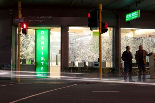 Nighttime street-level view of a commercial dental practice beneath a covered urban frontage. Large illuminated windows reveal a reception or waiting area, while a bright vertical green DENTISTS sign and green business sign identify the premises. Traffic lights, street signage, road markings, and blurred vehicle light trails occupy the foreground.