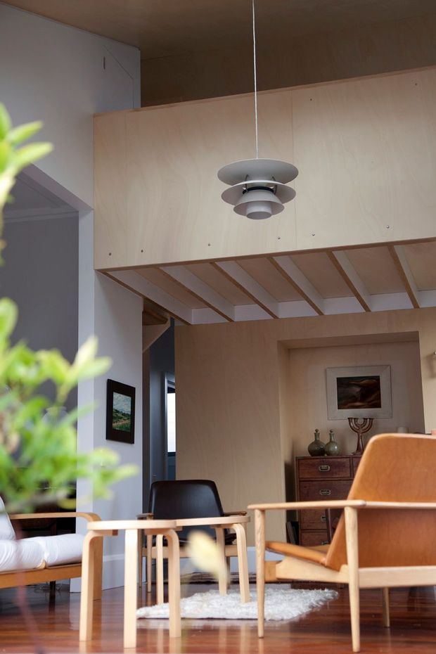 Warm, carefully composed interior view of a mid-century-inspired living room. Pale timber wall panelling and a timber-lined ceiling structure frame a suspended multi-tier pendant light. The room contains an upholstered lounge chair, dark armchair, low light-wood side table, dresser, artwork and decorative objects, with polished timber flooring and a small pale rug. A leafy plant partially obscures the foreground.