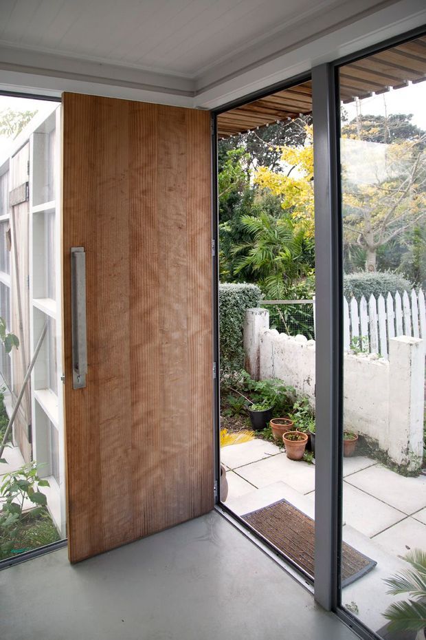 A bright contemporary entry area with a large pivot-style timber door and expansive dark-framed glass openings leading to a small paved garden courtyard. The door has vertically textured natural wood, while the interior features a smooth grey floor and a white panelled ceiling with simple cornice detailing. A timber pergola, planting, low masonry wall and white picket fence are visible outside.