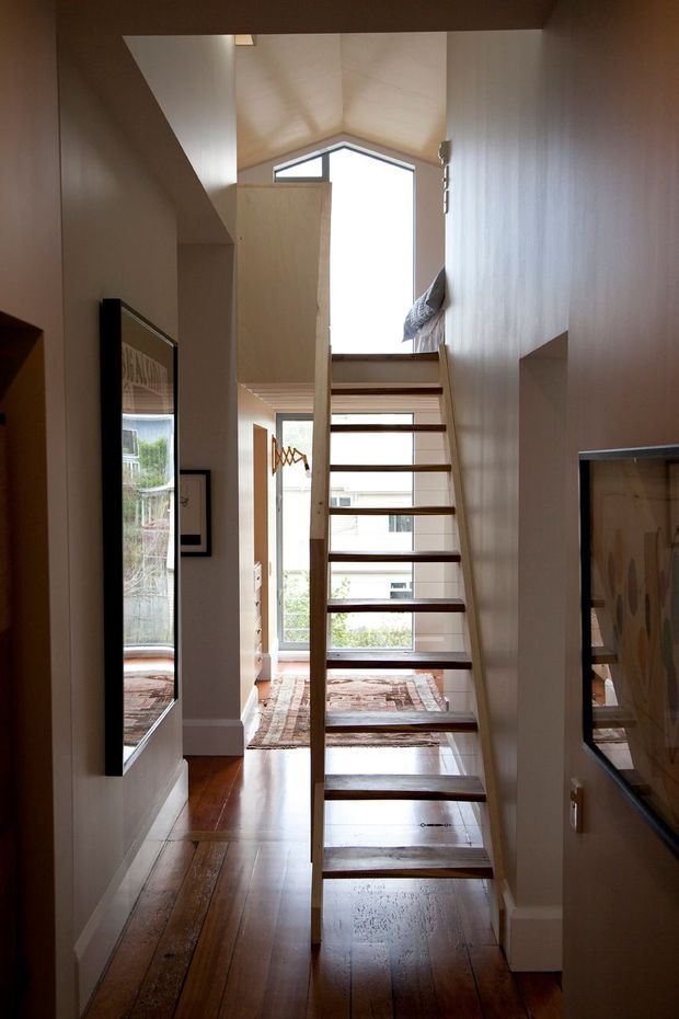 A narrow, light-filled interior stairway with an open-riser timber staircase leading to an upper level or loft. The stair is framed by pale timber-lined walls and ceiling, with polished dark timber flooring below, framed artwork on both sides, and large windows bringing in daylight. A distinctive gable-shaped clerestory window is visible above the landing.