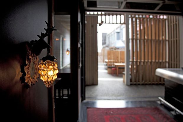 A dim, atmospheric interior passage opens through a large glazed doorway toward a small enclosed outdoor courtyard or patio. The foreground features a dark wall with an ornate hanging amber light and decorative branch-like wall element, while a red patterned rug leads toward the bright exterior. Vertical timber screening, exposed dark framing, and a reflective glass panel create a dramatic transition between inside and outside.