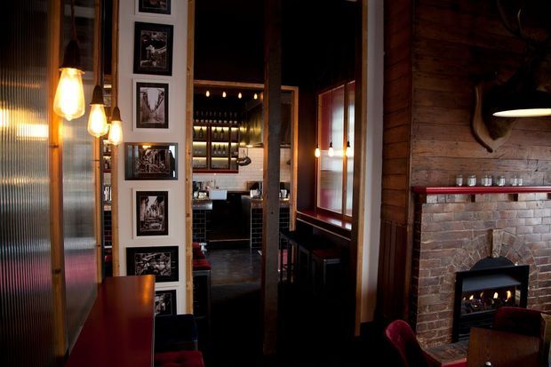 A dimly lit, intimate interior showing a rustic-industrial bar or dining space. The room features exposed timber wall cladding, a red-brown brick fireplace with a contemporary inset fire, dark timber floors and framing, translucent ribbed glazing, pendant lights, framed artwork, and a visible bar area with shelving and bottles in the background. Deep red upholstery and warm lighting create a moody, atmospheric setting.