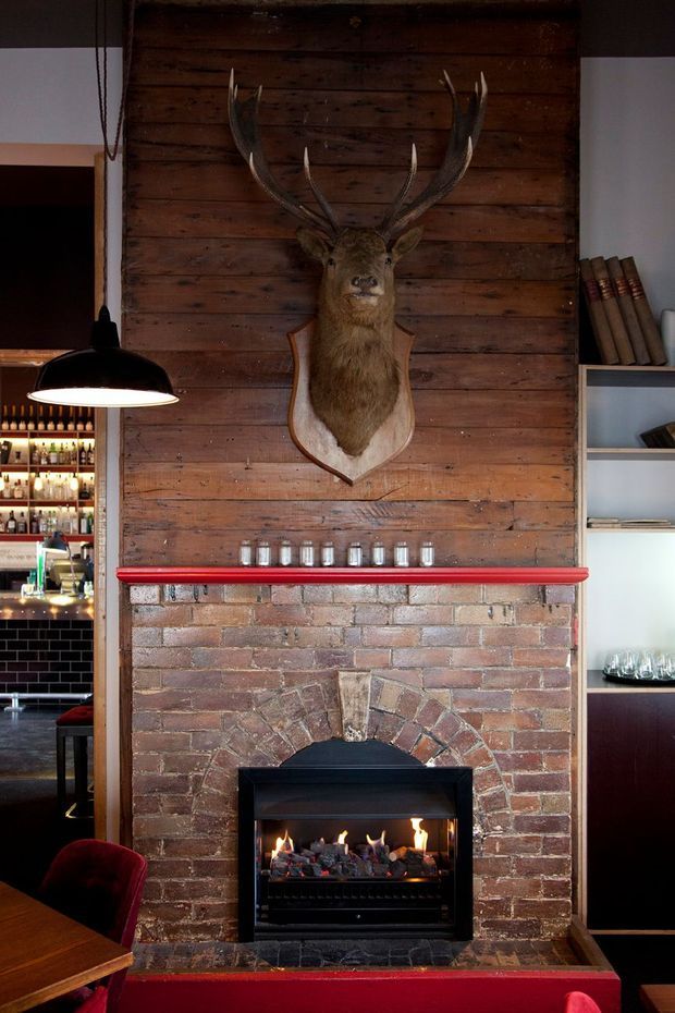 A warmly lit rustic hospitality interior centred on a substantial exposed brick fireplace with a dark timber mantel wall above it. A mounted deer head provides a strong feature, while open shelving, bottles, glassware and a black pendant light suggest a bar or restaurant setting. Deep red accents appear on the mantel trim and seating.