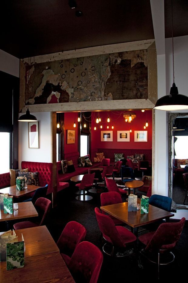 A dark, richly styled hospitality lounge or restaurant interior with numerous small timber dining tables, deep burgundy and blue upholstered chairs, tufted banquette seating, and a vivid red feature wall. Black pendant lights hang throughout the space, while exposed distressed wall surfaces and a large ornate mirror add a dramatic, eclectic character. The ceiling is dark and the floor appears dark carpet or another dark flooring finish.