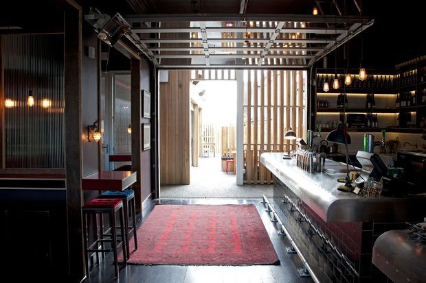 A dark, atmospheric commercial bar interior with an industrial fit-out. The space features a long stainless-steel bar counter, exposed metal framing, dark ribbed wall panels, suspended pendant bulbs, open shelving with bottles and glassware, bar stools, and a red patterned rug. A bright courtyard or exterior passage is visible beyond the bar.