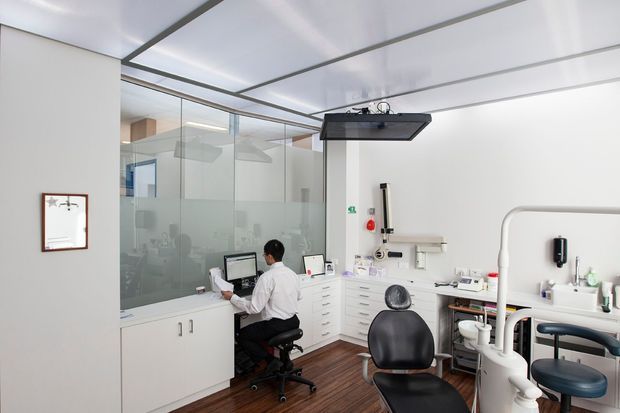 Bright modern dental treatment room with a dentist's workstation, dental chair and equipment, white built-in cabinetry, glass partitioning, and dark timber flooring. The room has a clean clinical fit-out with a suspended ceiling-mounted examination light and extensive dental fixtures.