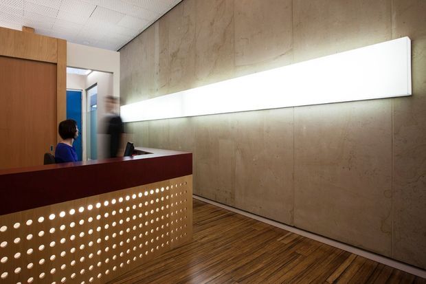 A contemporary reception area with a perforated wood reception desk featuring a dark reddish-brown top and fascia. Behind it, a long illuminated white wall panel is mounted on a textured concrete or plywood-like wall. The space has warm timber flooring, wood-clad joinery, and a suspended perforated acoustic ceiling. Staff and a computer are visible at the desk, indicating a commercial entry or reception setting.