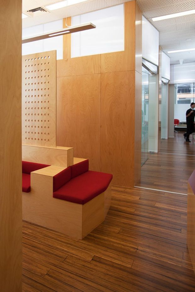 A modern commercial interior with warm timber wall panelling, matching timber flooring, and built-in angular lounge seating upholstered in deep red. A perforated timber screen adds a decorative architectural feature. The space opens toward a glazed corridor or adjacent office area, with linear suspended lighting and a perforated acoustic ceiling.