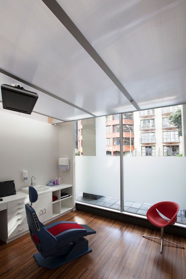 A bright modern dental or medical treatment room with a reclining navy and red chair, white built-in cabinetry and basin, wall-mounted equipment, a red guest chair, and large frosted-glass windows overlooking an urban apartment building. The room has a white paneled ceiling, pale walls, and warm timber flooring.