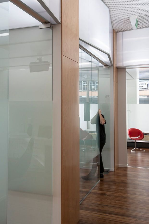 A bright contemporary commercial interior, likely an office, viewed along a glazed corridor or partitioned workspace. Full-height frosted and clear glass panels are framed by warm timber-clad structural elements, with dark timber flooring and a perforated white ceiling. A red modern lounge chair is visible in the adjoining space, while reflections show additional office furnishings and glazing. The composition emphasizes transparency, daylight, and clean architectural detailing.