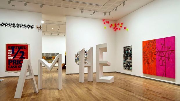 A bright contemporary art gallery interior with white walls, polished timber flooring, track lighting, and large colorful artworks displayed throughout. Sculptural white freestanding letter-like forms occupy the foreground, while framed and wall-mounted artworks add vivid red, pink, orange, and multicolored accents. A partial adjoining gallery space is visible beyond the opening.