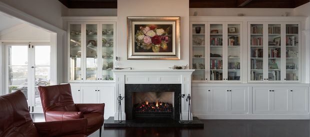 Traditional living room with a central white mantel fireplace, built-in white display and bookcase cabinetry, dark timber ceiling, dark flooring, and leather lounge chairs. Glazed doors and windows bring in natural light, while a framed floral artwork hangs above the fireplace.