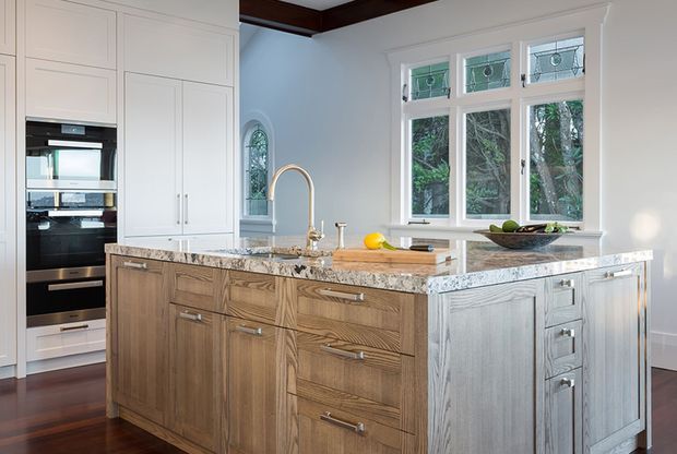 Bright, spacious traditional kitchen featuring a large central island with stained timber cabinetry, a white marble-look stone benchtop, integrated stainless-steel appliances, white perimeter cabinetry, dark timber flooring, and decorative leaded-glass windows. The island includes a tall gooseneck tap and generous drawer storage.