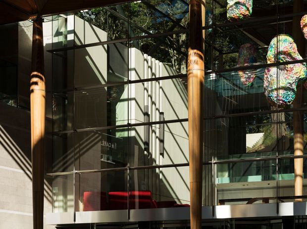 A dramatic contemporary interior atrium or public lobby with extensive floor-to-ceiling glazing, exposed structural framing, warm timber columns, strong geometric shadows, and suspended colourful sculptural light installations. A red seating area and upper-level circulation are visible behind the glass.