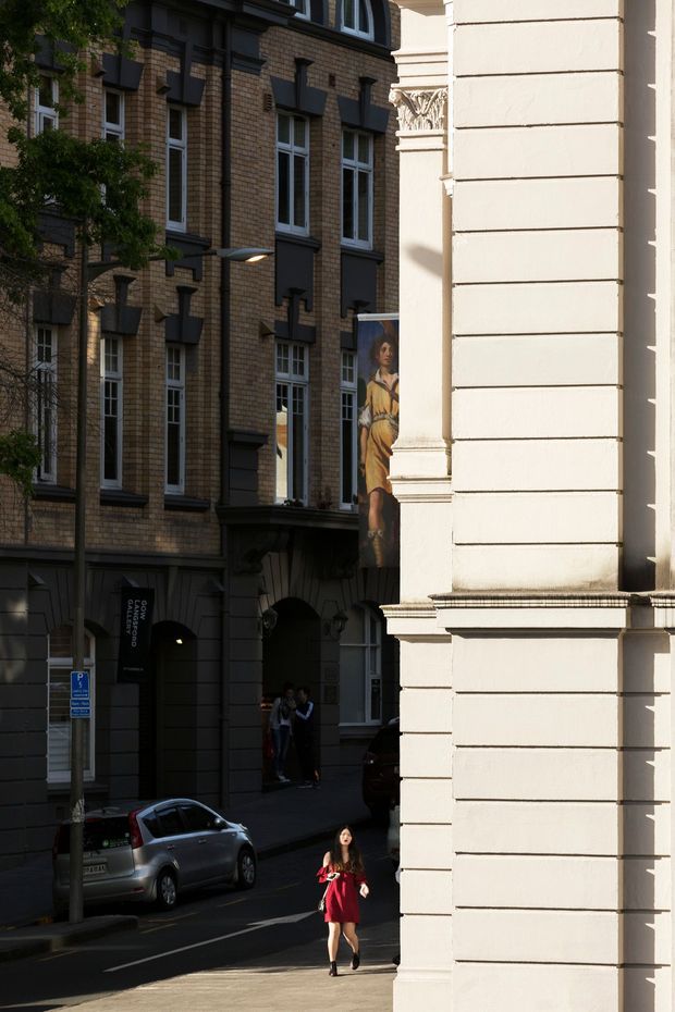 Street-level view of a dense urban commercial or civic streetscape. A large cream-painted masonry building with strong horizontal bands and a classical decorative capital dominates the right side, while the adjoining tan-brick building has repeated tall windows, dark projecting surrounds, arched entryways, and a covered passage. Cars, pedestrians, street lighting, and a mural/banner are visible in the shaded street.