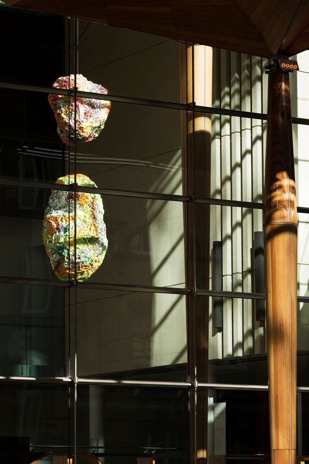 A tall, dramatic interior atrium with extensive floor-to-ceiling glazing and dark metal mullions, warm timber ceiling panels, and a prominent cylindrical timber-clad structural column. Two colourful sculptural pendant lights hang in front of the glazing, while strong sunlight creates geometric shadows across the walls and column. The image emphasizes the building’s vertical scale, structure, and layered reflections.