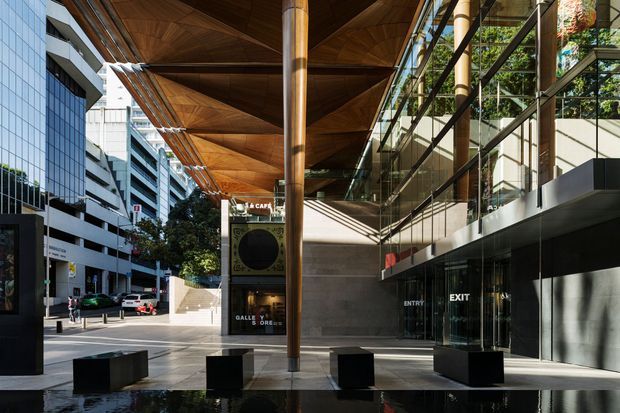 Urban commercial building entrance and plaza viewed from outside, featuring a dramatic angular timber canopy supported by a central column, extensive glass curtain walls, dark stone or concrete walls, reflective water, and integrated seating. The surrounding city streetscape and adjacent buildings are visible.