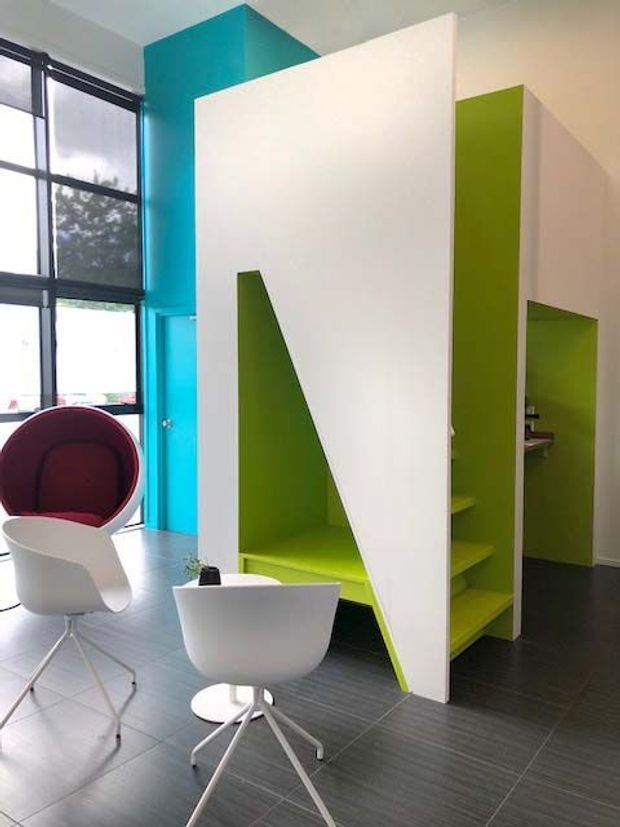 Bright contemporary interior with a striking turquoise wall, lime-green built-in angular shelving and partition elements, white geometric panels, dark grey tiled flooring, and floor-to-ceiling black-framed windows. Modern white swivel chairs and a red lounge chair furnish the open space.