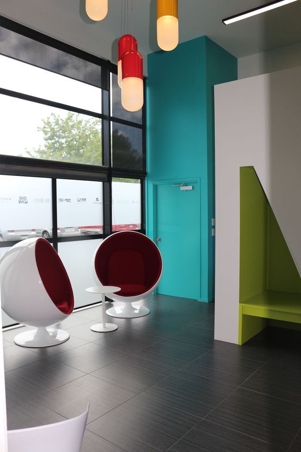 A bold contemporary commercial interior lobby or waiting area with floor-to-ceiling black-framed glazing, a vivid turquoise wall and door, dark tiled flooring, sculptural white-and-red egg chairs, and colorful pendant lights. A lime-green built-in bench or seating niche adds a geometric accent.