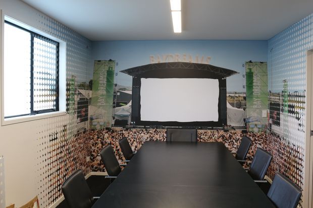 A compact conference or meeting room with a long black rectangular table and multiple black office chairs. The walls are covered with a large full-room mural depicting an outdoor concert or event stage and crowd, with cool blue-grey tones. A patterned privacy-glazed window is visible on the left, and a linear fluorescent ceiling light provides illumination.