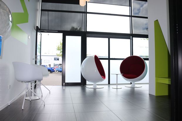 A bright contemporary commercial-style lobby or waiting area with expansive black-framed floor-to-ceiling glazing, a glazed entrance door, large grey tiled flooring, white sculptural chairs, and distinctive white-and-red spherical lounge chairs. Lime-green geometric wall graphics and cabinetry add a bold accent, while daylight creates an airy, polished atmosphere.