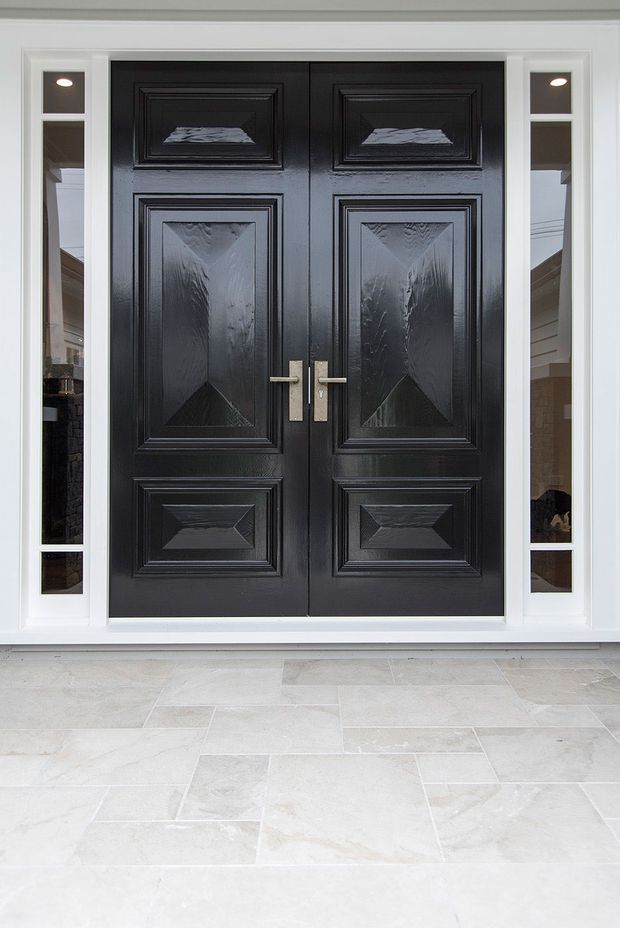 Front-facing view of a formal residential entry with a pair of black raised-panel timber doors, brushed metal handles, white surrounding trim and narrow sidelights. The entrance opens onto a pale large-format tiled or stone-paved floor, with limited interior and exterior context visible through the glazing.