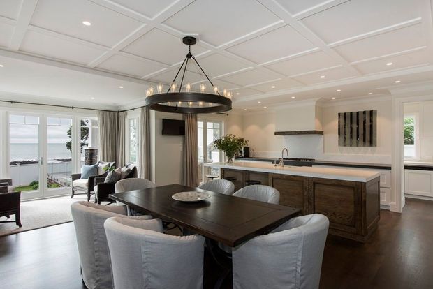 Bright open-plan dining and kitchen interior with a large dark timber dining table surrounded by slipcovered chairs, a substantial kitchen island, white cabinetry, dark timber flooring, and a coffered white ceiling. Large glazed doors and windows open toward a waterfront view. A statement circular pendant light anchors the dining area.