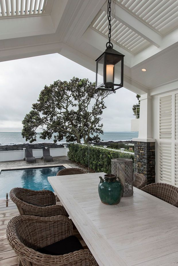A covered coastal outdoor dining terrace with a large whitewashed dining table and woven wicker chairs beside a rectangular swimming pool. White architectural columns, louvred shutters, a slatted pergola ceiling, and a hanging lantern frame views toward the ocean and rocky shoreline. Stone-clad columns and clipped hedging add texture, while a mature coastal tree and subdued blue-grey palette create a refined resort-like atmosphere.