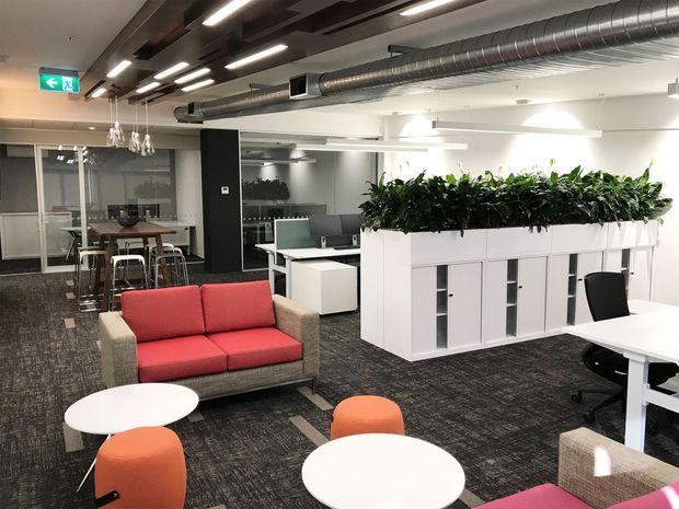 Open-plan contemporary office interior with exposed silver HVAC ductwork, linear ceiling lighting, dark carpet flooring, white built-in storage planters, workstations, meeting tables, and a red sofa with orange lounge chairs. Glazed meeting rooms are visible in the background.