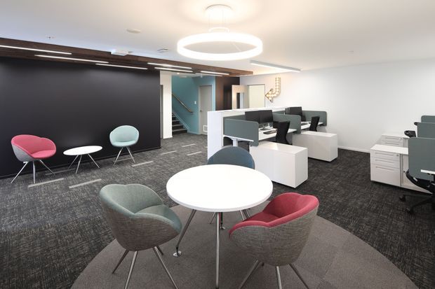 A bright, contemporary open-plan commercial office with shared workstations, meeting tables, task seating, a dark feature wall, patterned carpet, and distinctive circular pendant lighting. A small stair and teal-painted circulation area are visible toward the rear.