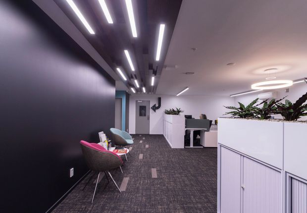 A contemporary commercial office interior with an open-plan workspace, reception or waiting chairs, white partitioned workstations with integrated planter boxes, dark carpet tiles, and a striking linear LED ceiling-light installation. The space has a monochromatic palette with black, white, grey, and muted purple tones.