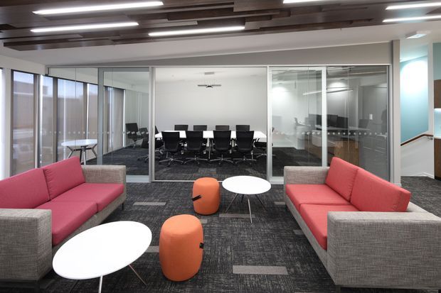 Contemporary commercial office interior with a relaxed breakout lounge in the foreground, furnished with coral-red upholstered sofas, orange ottomans, and white oval coffee tables. A glazed meeting room and additional glass-partitioned workspace are visible behind, with dark patterned carpet, exposed timber ceiling battens, and linear recessed lighting.