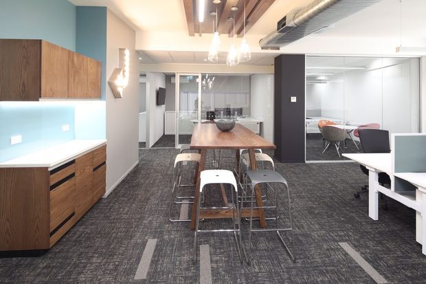 Open-plan contemporary commercial office interior with a central timber communal dining or breakout table, stools, carpet tile flooring, a kitchenette with dark timber cabinetry and white benchtop, glass-partitioned meeting spaces, and white workstations. Exposed timber ceiling battens and visible HVAC ductwork add architectural interest.