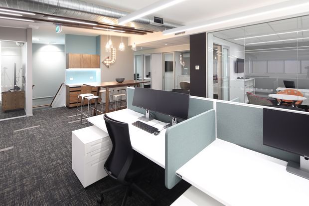 A bright contemporary open-plan office with white workstations, pale blue privacy screens, black ergonomic chairs, and computer monitors. A communal kitchenette or break area with timber cabinetry, stools, pendant lighting, and a small dining table sits toward the rear. Glass-partitioned meeting rooms, exposed ductwork, carpet tiles, and linear ceiling lighting contribute to a modern commercial fit-out.