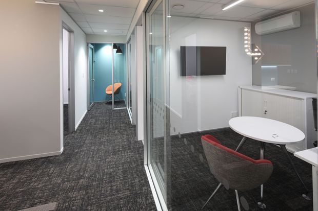 Contemporary commercial office interior viewed from a carpeted corridor, with a glazed meeting room containing an oval white table and upholstered chairs. The space features white walls, dark patterned carpet, glass partitions, built-in cabinetry, a wall-mounted display, recessed lighting, and an air-conditioning unit.
