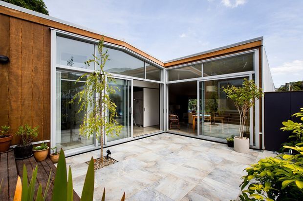 Modern single-storey courtyard house with a compact L-shaped form, warm timber-clad walls, expansive aluminium-framed glazing, and a paved internal courtyard. Interior dining and living areas are visible through the glass, with established planting around the courtyard edges.