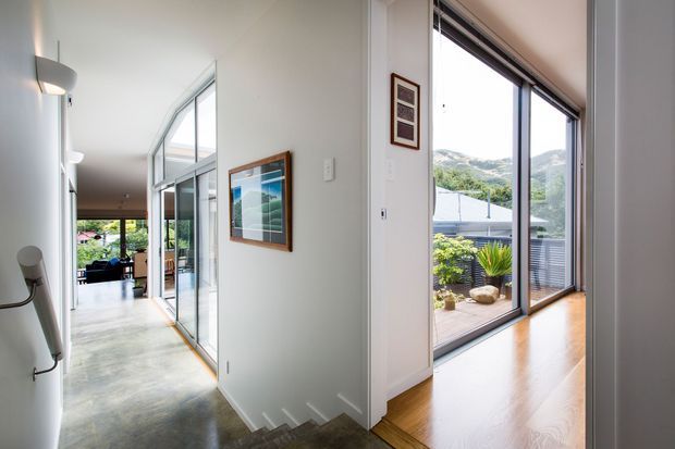 Bright contemporary interior hallway and stair landing with polished concrete flooring, white walls, extensive floor-to-ceiling glazing and timber-framed sliding doors. The corridor opens toward a living area and landscaped outdoor space, while a stair descends in the foreground. Warm timber flooring is visible in the adjacent room, with mountain scenery and a terrace seen through the glazing.