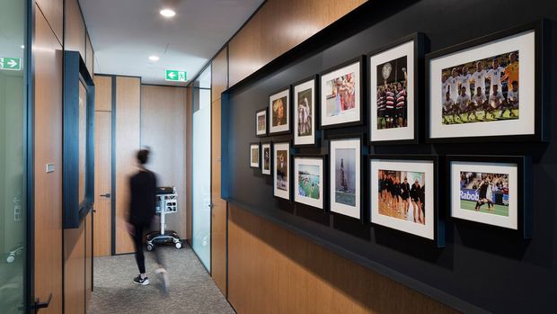 A contemporary interior corridor or hallway with dark charcoal feature walls, warm timber wall panelling, recessed ceiling lighting, and framed sports photography arranged in a gallery display. Light timber doors and panels line the passage, with emergency exit signage and a cleaning trolley visible in the distance.